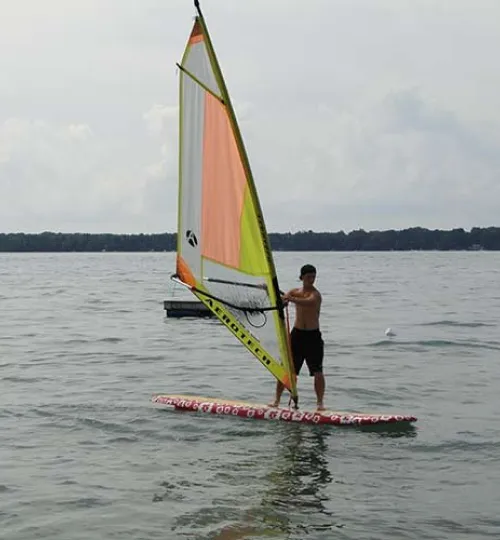A person stands on a windsurfing board in calm water at Torch Lake, holding the sail upright. The sky is cloudy and a forested shoreline is visible in the background, perfect for guests staying at a nearby bed and breakfast.