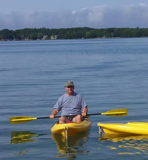 A man in a gray shirt and cap paddles a yellow kayak on calm Torch Lake, with trees and houses—perhaps a cozy bed and breakfast—on the distant shore under a partly cloudy sky. Another yellow kayak floats nearby.