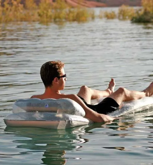 A man wearing sunglasses and black swim trunks relaxes on a transparent inflatable mattress, floating on the calm waters of Torch Lake with greenery and sandy hills in the background, capturing the perfect bed and breakfast getaway.