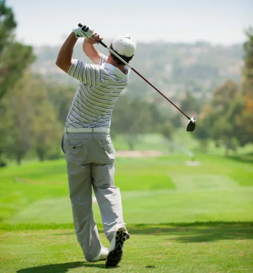 A golfer in a striped shirt and light pants finishes a swing on a lush green golf course near Torch Lake, facing away from the camera toward the fairway and distant trees, steps from a charming local bed and breakfast.