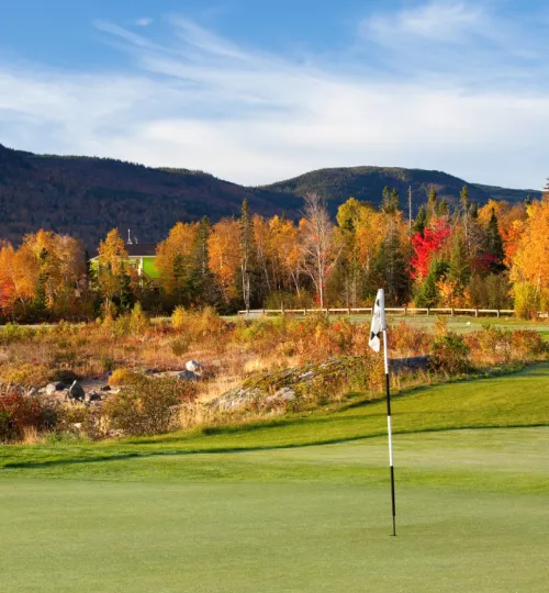 A golf flag stands on a green, surrounded by trees with vibrant autumn foliage, with hills and a blue sky in the background—just steps from a charming Torch Lake bed and breakfast.