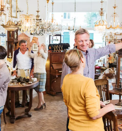 People browse antiques and vintage items in a well-lit shop filled with chandeliers, furniture, and decorative pieces—perfect treasures for a cozy Torch Lake bed and breakfast. A man smiles and gestures while talking to a woman in the foreground.