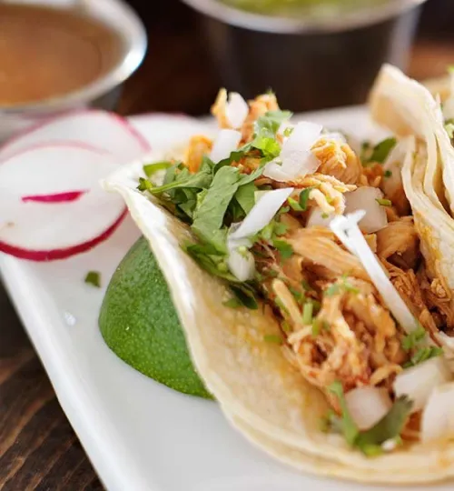 Close-up of a chicken taco in a corn tortilla, topped with onions and cilantro, served with lime wedge and sliced radishes on a white plate—perfect for enjoying at a cozy Torch Lake bed and breakfast. A cup of sauce is blurred in the background.