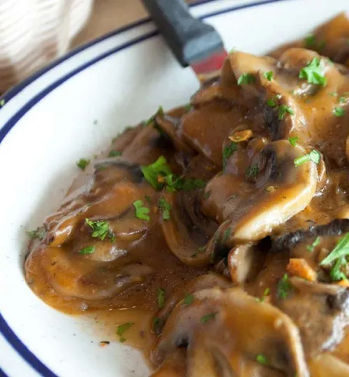 Close-up of ravioli smothered in a brown mushroom sauce, garnished with chopped parsley, served on a white plate with a blue rim. This delicious dish is a guest favorite at our Torch Lake bed and breakfast. A fork is partially visible in the background.