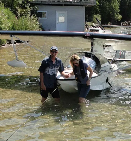 Two people stand in shallow water beside a small seaplane on Torch Lake, smiling. The seaplane rests near a shore with a cozy bed and breakfast, trees, and a dock visible in the background.