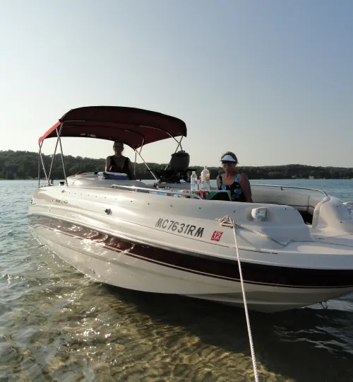 Two people relax on a motorboat anchored in shallow, clear water near the tree-lined shoreline of Torch Lake. The boat, with a red canopy and supplies on deck, is perfect for guests from a nearby bed and breakfast seeking a peaceful getaway.