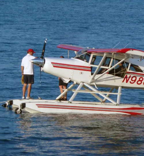 A man in a white shirt and red cap stands on the float of a red and white seaplane on Torch Lake. Another person is partially visible near the plane’s cockpit, possibly arriving at or departing from a nearby bed and breakfast.