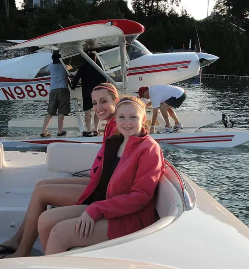 Two smiling young women sit on a boat in the foreground, enjoying a sunny day on Torch Lake. Behind them, two people are working on a red-and-white seaplane floating near the tree-lined shore, perfect for those staying at a nearby bed and breakfast.