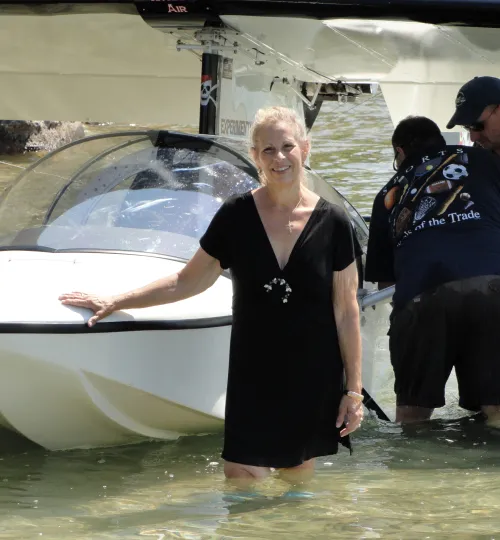 A smiling woman in a black dress stands in shallow Torch Lake water, resting her hand on a small white boat with a glass canopy. Another person is beside the boat, partially turned away and wearing a cap, near a charming bed and breakfast.