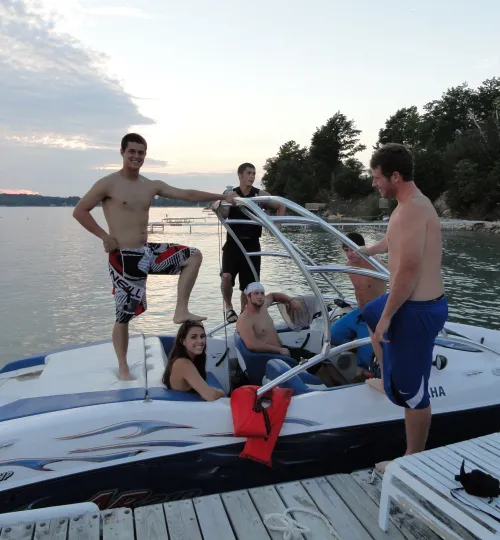 A group of young adults, some shirtless, relax and pose on a speedboat docked by Torch Lake. Trees and calm water are in the background as they enjoy a summer day outdoors, perhaps after staying at a cozy bed and breakfast nearby.