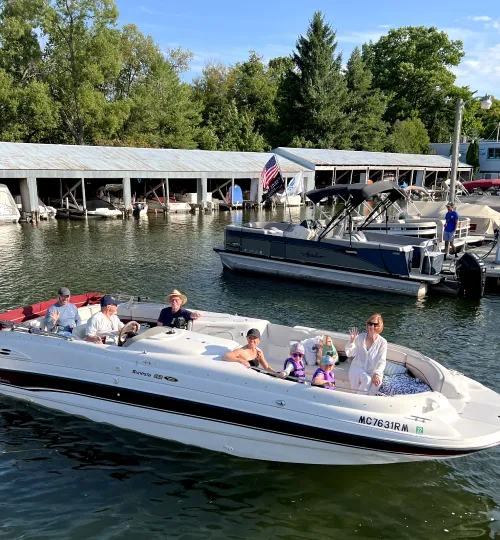 A group of six people, including two children, relax on a white speedboat near a Torch Lake marina on a sunny day, with a bed and breakfast, another pontoon boat, and trees visible in the background.