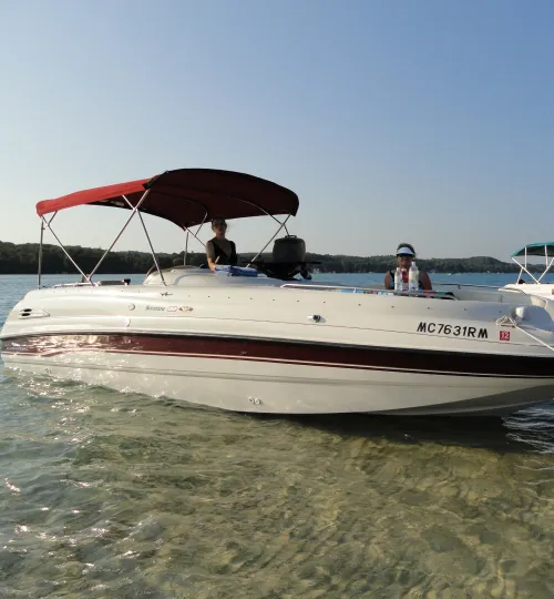 A white and red motorboat with a maroon canopy floats in the clear, shallow waters of Torch Lake near the shoreline. Two people enjoy a sunny day on board, making memories near one of the area’s charming bed and breakfast spots.
