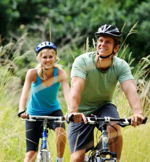 A man and a woman wearing helmets ride bicycles on a grassy trail near Torch Lake, smiling and enjoying the outdoors with green trees in the background, perhaps heading back to their cozy bed and breakfast.