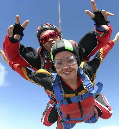 Two people skydiving in tandem, both wearing helmets and goggles, with their arms spread out. The person in front is smiling while the instructor behind flashes a peace sign against a clear blue sky above scenic Torch Lake.
