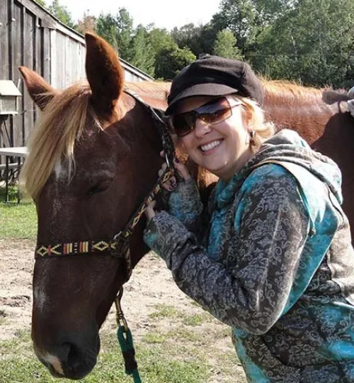 A smiling woman in sunglasses and a cap hugs a brown horse outdoors near a wooden building at a bed and breakfast, with green trees and hints of Torch Lake in the background.