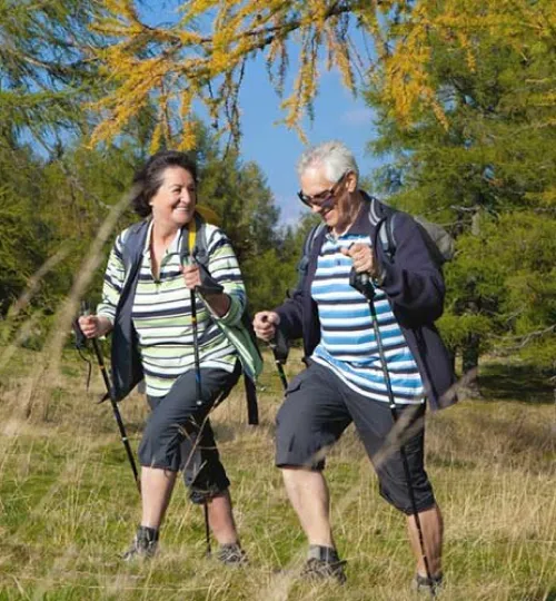 Two older adults, one with dark hair and one with white hair, are smiling and using walking poles while hiking through a grassy, wooded area near Torch Lake on a sunny day—perhaps exploring trails from their cozy bed and breakfast.