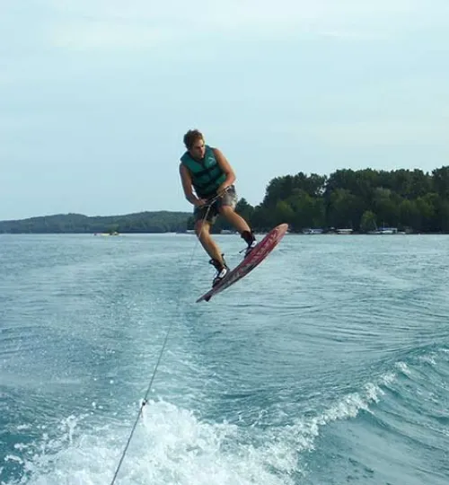 A person wearing a green life vest is wakeboarding mid-air above Torch Lake, holding onto a tow rope, with trees and shoreline—including a charming bed and breakfast—visible in the background.