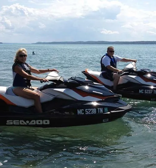Two people wearing life jackets ride black and white Sea-Doo jet skis on calm Torch Lake, with distant land and a cloudy sky in the background—perfect for guests staying at a nearby bed and breakfast.
