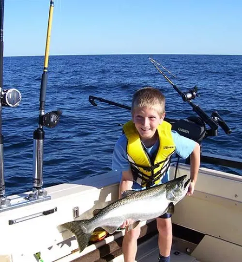 A smiling boy wearing a yellow life jacket holds a large fish on a boat at Torch Lake, with fishing rods and the open blue sea in the background under a clear sky—perfect for a stay at a nearby bed and breakfast.