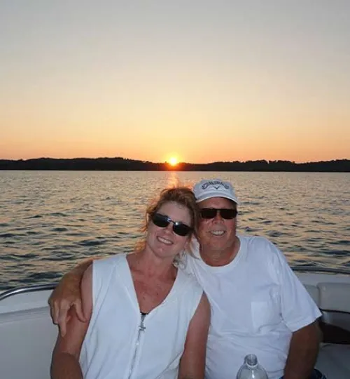 A smiling couple wearing sunglasses sits together on a boat at Torch Lake, the man's arm around the woman. The sun is setting over calm water and a distant shoreline, capping off a perfect day from their cozy bed and breakfast.