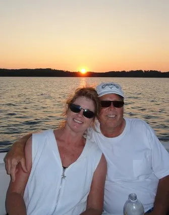 A smiling couple wearing sunglasses and white tops sit together on a boat at sunset on Torch Lake, with calm water and a low sun on the horizon—perfect for a bed and breakfast getaway.