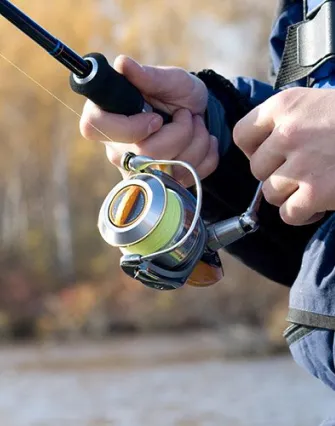 Close-up of a person holding and using a fishing rod and reel, wearing a blue jacket, with Torch Lake’s tranquil waters and trees blurred in the background—perfect for a relaxing getaway at a nearby bed and breakfast.