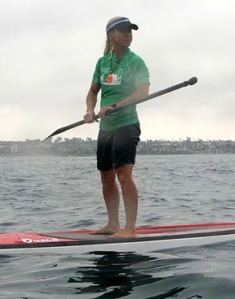 A person wearing a green shirt, black shorts, and a cap is standing on a paddleboard in Torch Lake, holding a paddle. The sky is cloudy, and a bed and breakfast can be seen along the visible shoreline in the background.