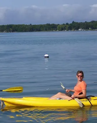 A woman wearing sunglasses and an orange shirt smiles while paddling a yellow kayak on calm Torch Lake, with trees, a buoy, and a charming bed and breakfast in the background.