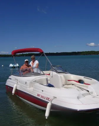 Two people are sitting in a white and red motorboat on Torch Lake under a clear blue sky, with trees visible in the background—a perfect scene for a relaxing stay at a nearby bed and breakfast.
