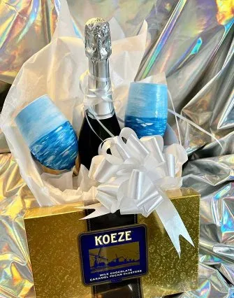 A gift set with a gold Koeze chocolate box, sparkling wine, and two hand-painted blue wine glasses—perfect for enjoying at a cozy bed and breakfast or by the serene shores of Torch Lake—all wrapped in white tissue with a large ribbon.