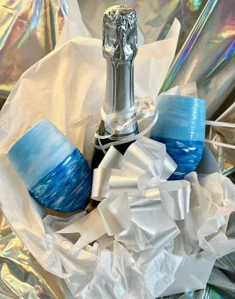 A bottle of champagne with a silver foil top is nestled in white tissue paper beside two blue ombré wine glasses and a large white ribbon bow, perfect for a cozy evening at a Torch Lake bed and breakfast, all set against an iridescent background.