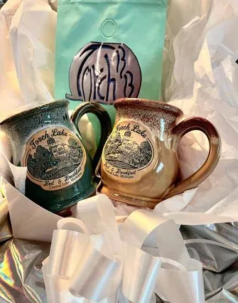 Two ceramic mugs labeled "Torch Lake Bed & Breakfast" rest on crumpled white tissue paper beside a teal coffee bag with a silver logo. White ribbon and shiny silver fabric add to the charming bed and breakfast gift presentation.
