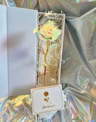 A gold-stemmed iridescent artificial rose rests in a white gift box lined with shredded paper. In front is a card with a rose illustration and "Just for you"—a perfect keepsake from your stay at a cozy Torch Lake bed and breakfast.