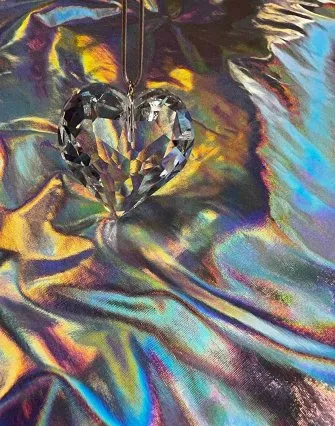 A faceted crystal heart pendant hangs above an iridescent, rainbow-colored fabric that reflects light in shimmering shades—reminiscent of the tranquil blues and purples of Torch Lake, perfect for a cozy bed and breakfast setting.