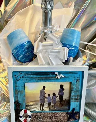 A gift set featuring a champagne bottle, two blue ombre glasses, a white bow, and a beach-themed photo frame with seashells and a family photo—perfect for reliving cherished Torch Lake or bed and breakfast memories—all set against metallic wrapping.