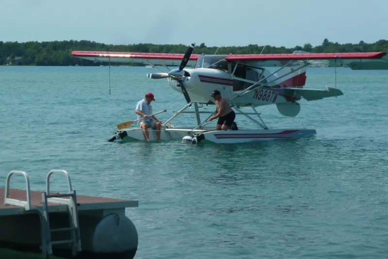 Two people enjoy sitting on the floats of a floatplane in Torch Lake near a dock; one paddles while the other relaxes, with a forested shore behind them under a clear sky—an idyllic scene near a cozy bed and breakfast.