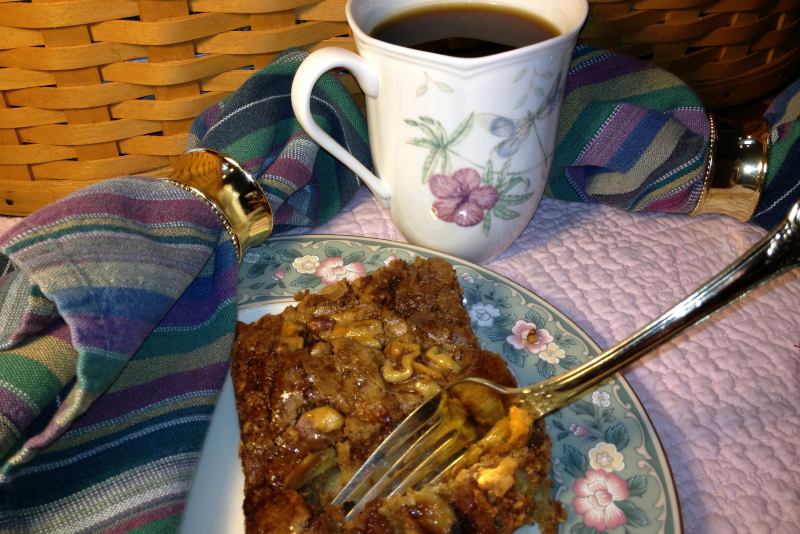 A floral-patterned plate with a slice of nutty dessert and a fork sits beside a mug of coffee at a cozy bed and breakfast, with a striped cloth napkin and gold ring on a textured surface, set near a wicker basket.