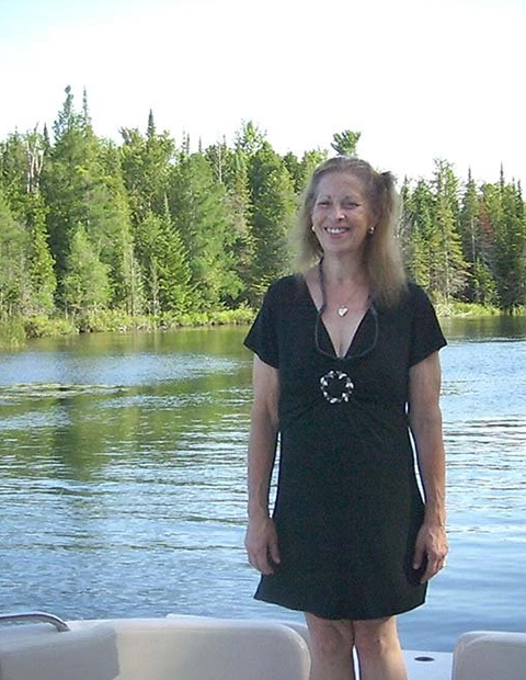 A woman in a black dress stands on a boat on Torch Lake, smiling in front of calm waters and green trees under a clear sky. The pleasant weather hints at the perfect day for guests staying at a nearby bed and breakfast.