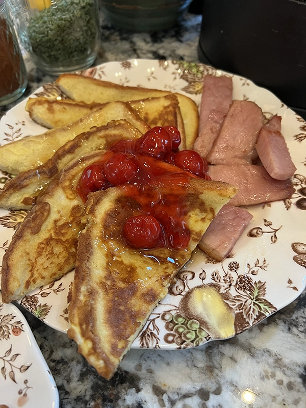 A floral-patterned plate with slices of French toast topped with cherry sauce, served alongside pieces of ham on a granite countertop—perfect for a cozy morning at a Torch Lake bed and breakfast.