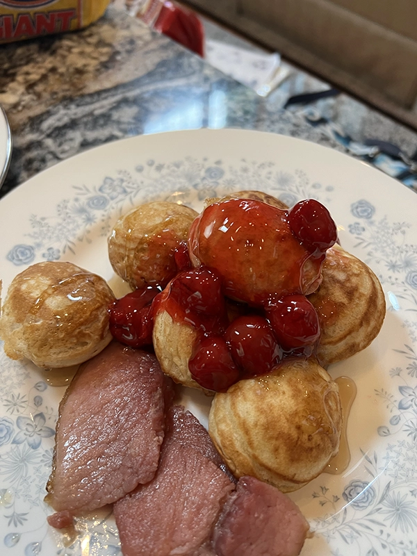 A plate with three slices of ham and several Danish aebleskiver pancakes topped with cherries and syrup, served on a white floral-patterned dish—perfect for a cozy bed and breakfast near Torch Lake.