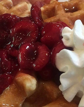 A close-up of a waffle topped with glossy cherry compote and a swirl of whipped cream, perfect for a cozy morning at a bed and breakfast near scenic Torch Lake.