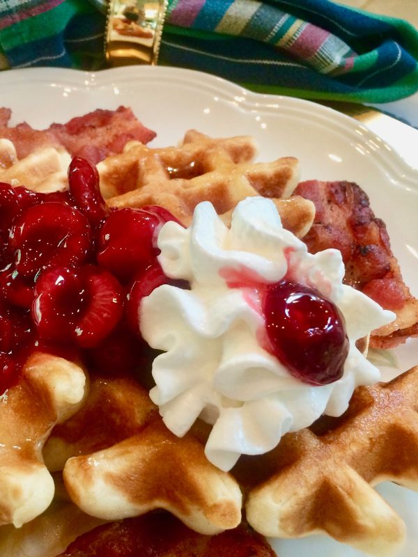 A close-up of waffles topped with whipped cream and cherry sauce, served with strips of bacon on a white plate. A colorful napkin with a gold napkin ring is in the background.